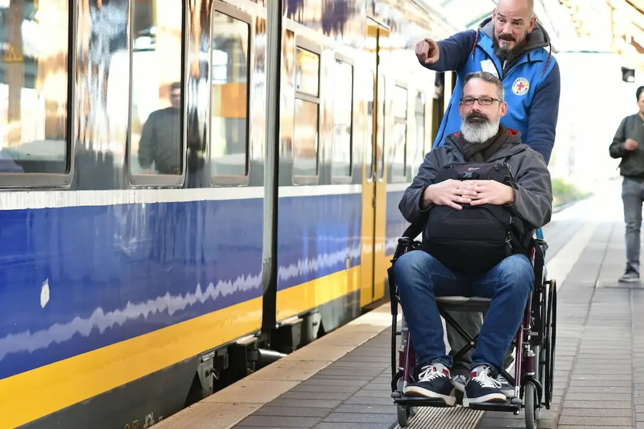 Ein Mitarbeiter der Bahnhofsmission und Rollstuhlfahrer mit Rucksack stehen am Bahnhof neben einem Zug. Der Mitarbeiter der Bahnhofsmission zeigt mit der Hand in Richtung Zug. Eine weitere, unkenntliche Person im Hintergrund.