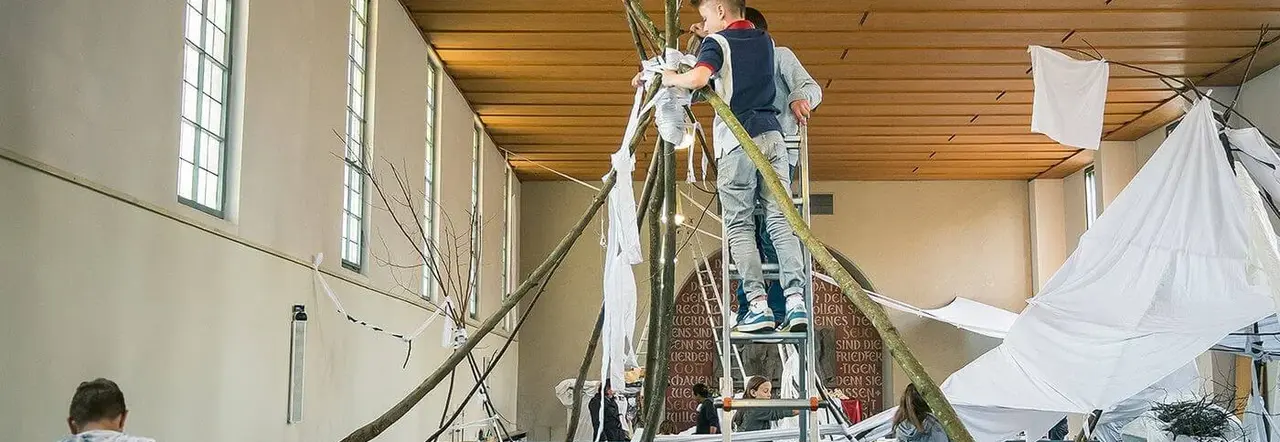 Im Kirchenraum hat sich eine große zeltartige Skulptur aus Ästen und weißen Stoffen entwickelt. Thomas Putze hat die Materialien bereits im Vorfeld aus den Gärten und Schränken vieler Gemeindeglieder bekommen. Im Kirchenraum verbindet sich dieses Material – ein starkes Symbol für Kirche.